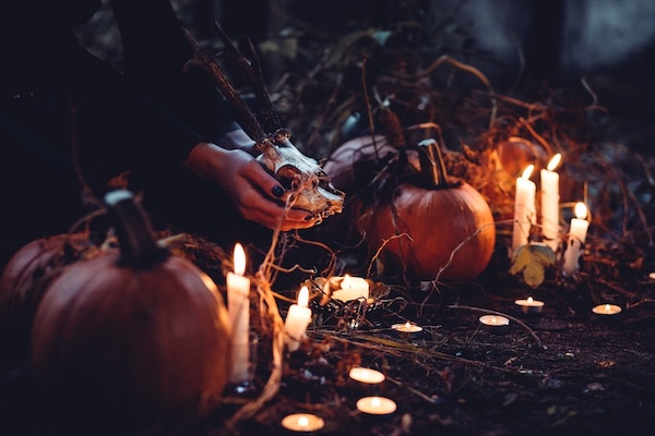 On a night-time woodland floor can be seen a variety of lit tealights and candles, along with some pumpkins. A black-clad woman's arms holds a deer's skull over one of the candles.